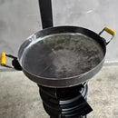 Chapa de ferro com alças amarelas sobre fogareiro portátil preto em fundo cinza