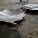 Panelas grandes de metal com alças de madeira, formato de folha, sobre chão de cimento.